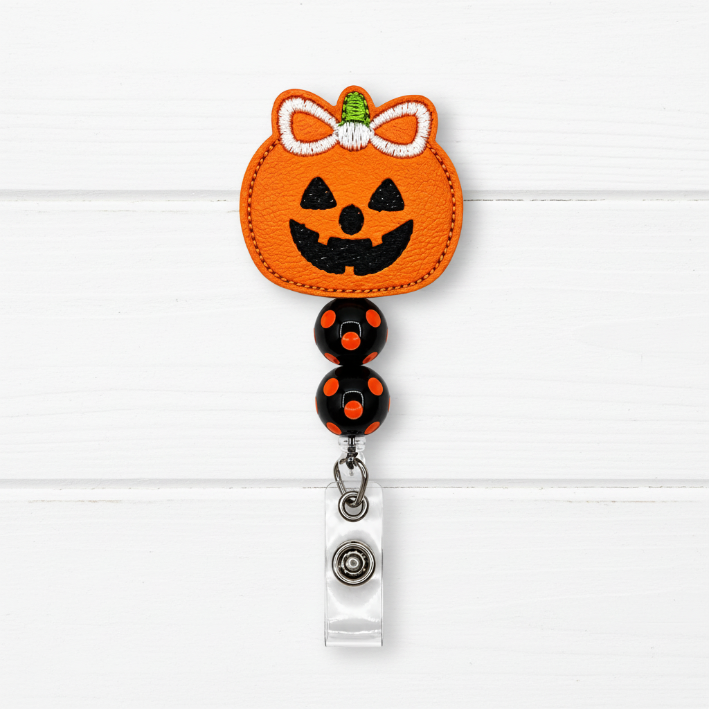 A Halloween-themed retractable badge reel featuring an orange pumpkin patch with a black jack-o'-lantern face, white bow, and green stem, attached to black beads with orange dots and a silver clip on a white wood plank background.