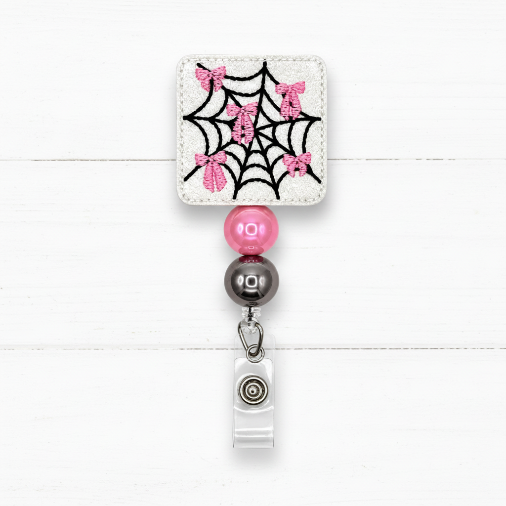 A square embroidered patch with a black spiderweb and pink glitter bows on a white fabric background, attached to a retractable badge reel with pink and dark gray beads.