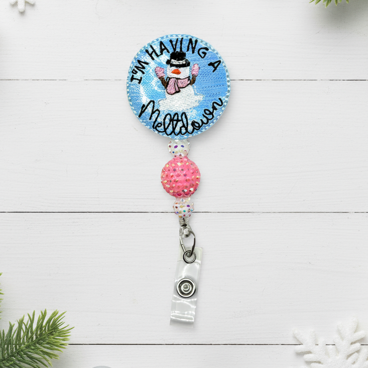 A badge reel with a circular silver and blue clip, and a decorative phrase that reads 'I'm Having A Meltdown' with a small snowman figure.