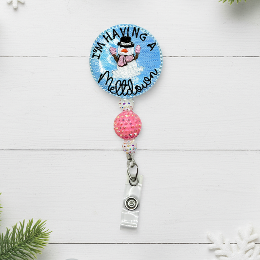 A badge reel with a circular silver and blue clip, and a decorative phrase that reads 'I'm Having A Meltdown' with a small snowman figure.