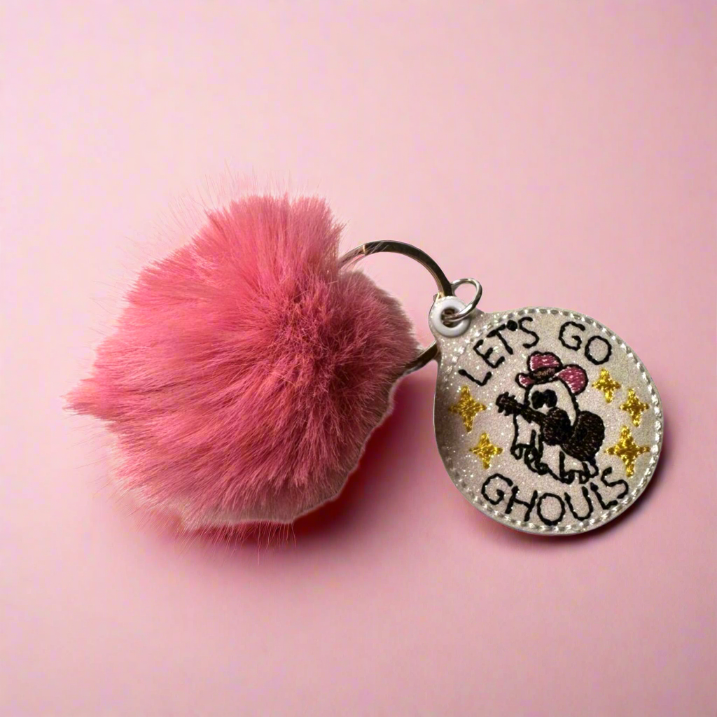 A pink pom pom keychain with a circular keyring and a charm featuring the text 'LETS GO GHOULS' on a pink background.