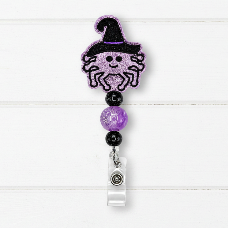 A Halloween-themed retractable badge reel featuring a glittery purple spider wearing a black witch hat, separated by purple and black beads from a clear plastic clip on a white wood plank background.