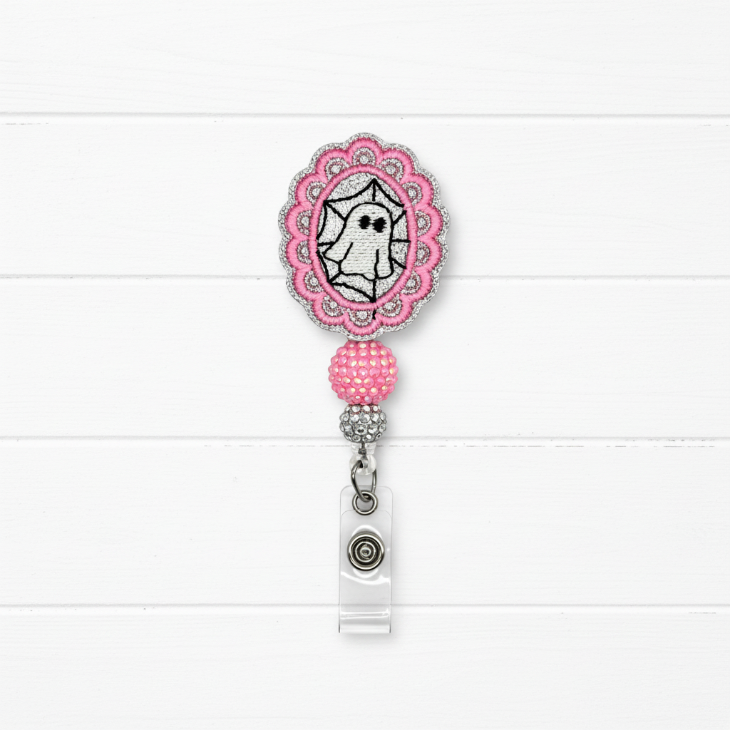 A pink and silver embroidered badge reel with a ghost design, shown on a white wood plank background.
