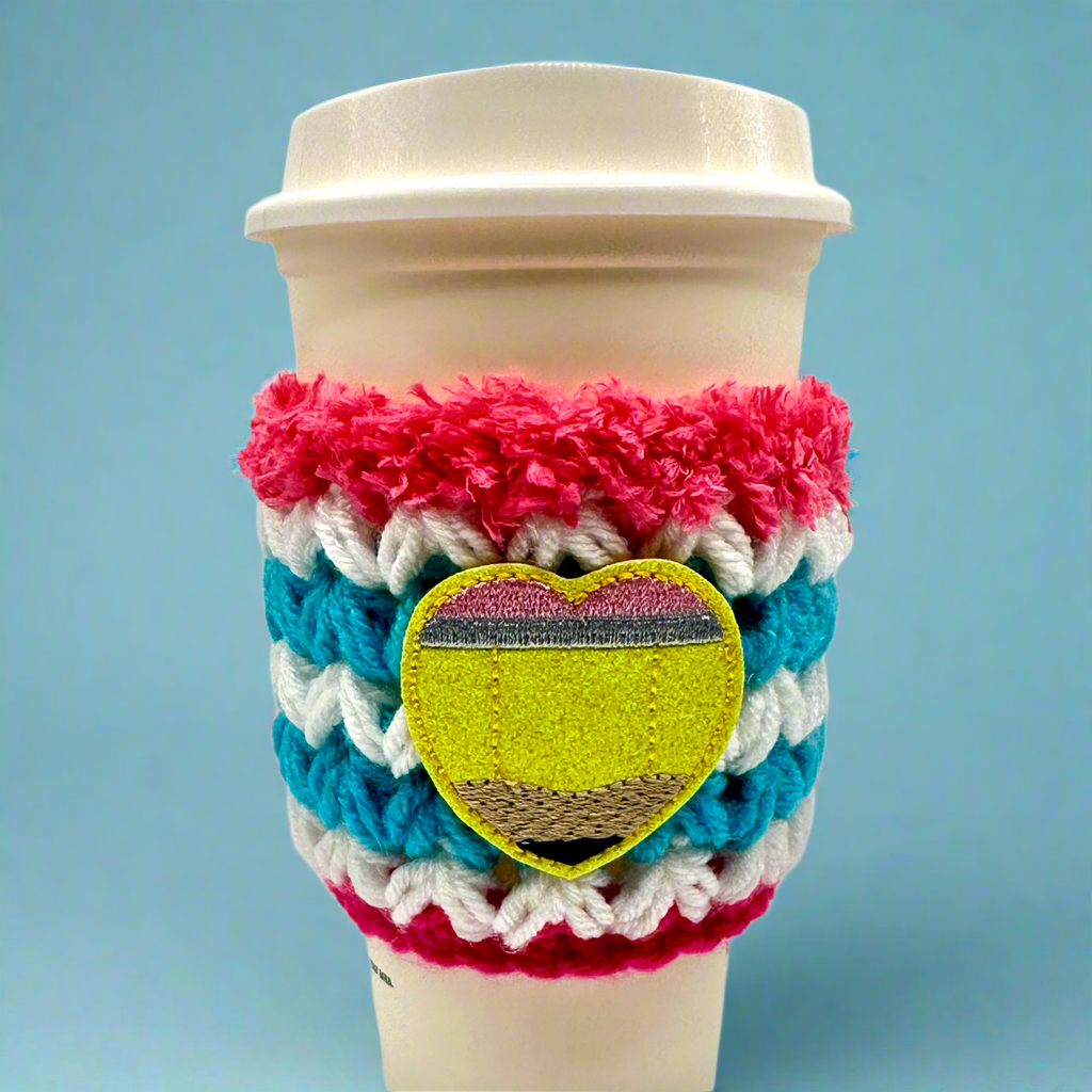 A crocheted coffee cozy featuring a geometric pattern with a heart shape in the center, in colors blue, pink, white, and yellow, displayed on a white coffee cup.