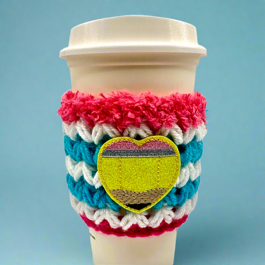 A crocheted coffee cozy featuring a geometric pattern with a heart shape in the center, in colors blue, pink, white, and yellow, displayed on a white coffee cup.