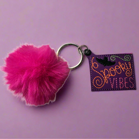 A fuchsia pink pom pom keychain with a black strap featuring the text 'Spooky Vibes' on a purple background.