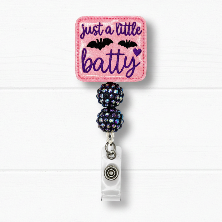 A pink embroidered badge reel with the text 'just a little batty' in purple script, decorated with two black bats and a small purple heart. The pink square patch is attached to two dark iridescent rhinestone-covered beads and a silver metal clip, set against a white wood plank background.