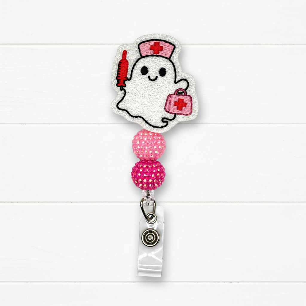 A ghost-shaped embroidered badge reel cover featuring a white ghost with a red nurse hat, red syringe, and pink first-aid kit, attached to a retractable clip with pink rhinestone beads on a white wood background.