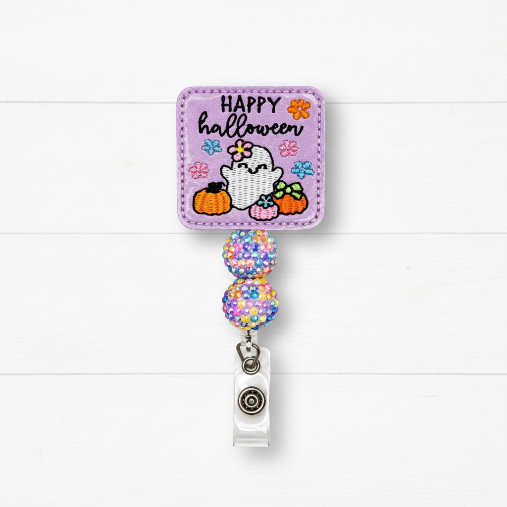 A Halloween-themed retractable badge reel with a lavender embroidered patch featuring a ghost, pumpkins, flowers, and the text 'HAPPY halloween.' The patch is attached to two glittery multicolor beads and a clear plastic clip with a silver metal snap, shown against a white wood plank background.