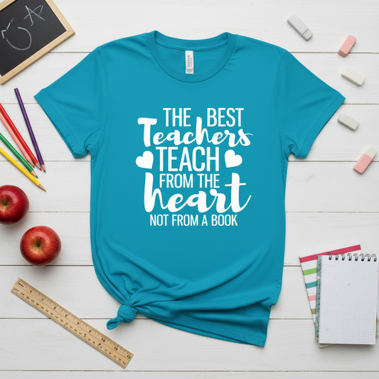 Blue t-shirt with 'The Best Teachers Teach from the Heart Not from a Book' text on a wooden surface with school supplies.