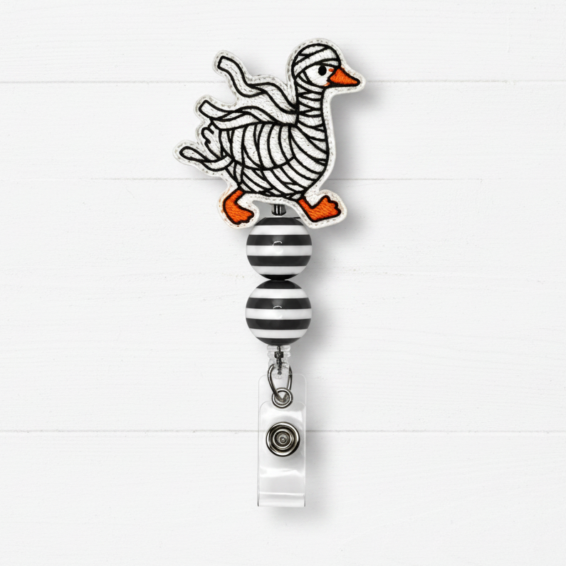A novelty retractable badge reel featuring a whimsical cartoon duck with black-and-white striped body bands and bright orange feet and beak, mounted above a black-and-white striped roller ball and white clip on a white wood plank background.