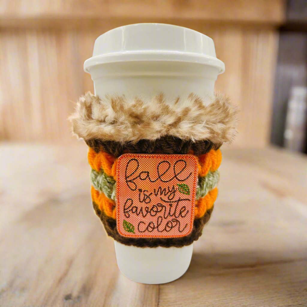 Coffee cup with a cozy holder and 'Fall is my favorite color' patch on a wooden surface.