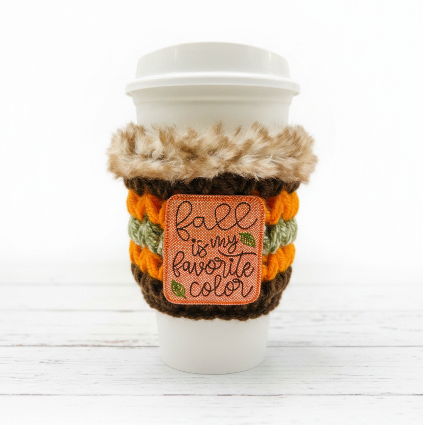 Coffee cup with a cozy holder and 'Fall is my favorite color' patch on a wooden surface.