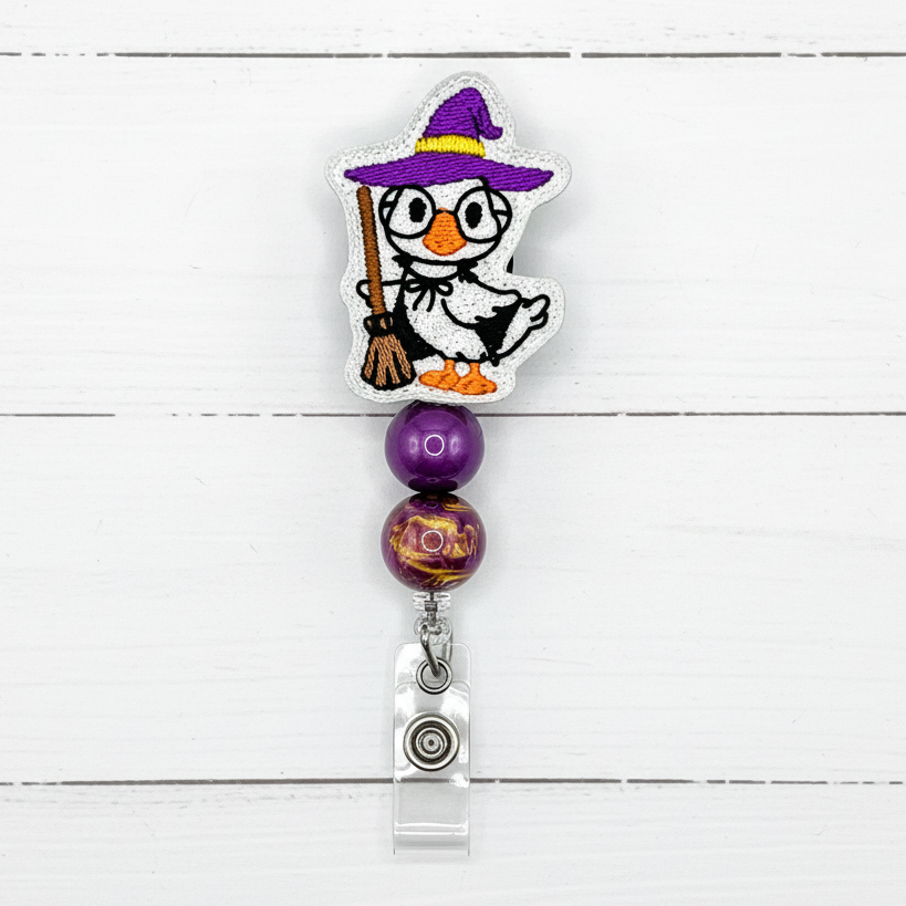 A Halloween-themed retractable badge reel featuring a white penguin dressed as a witch with a purple hat and broom, two purple beads, and a silver metal clip on a white wood plank background.