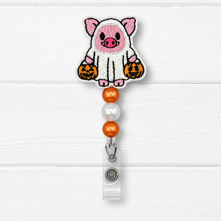 A Halloween-themed embroidered badge reel clip featuring a cute pink pig dressed in a white ghost with orange jack-o'-lanterns, hanging from orange and pearl beads on a white wood plank background.