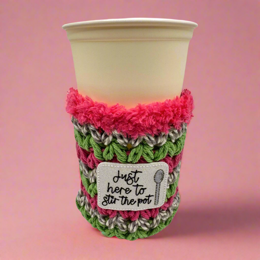 A crocheted multicolored cup cozy with text 'Just here to stir the pot' displayed on a beige cup.