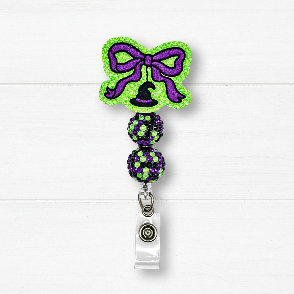 Halloween-themed retractable badge reel with a green glitter bow, purple glitter accents, a black witch hat, and two glittery purple and green beads on a clear plastic clip, shown on a white wood background.