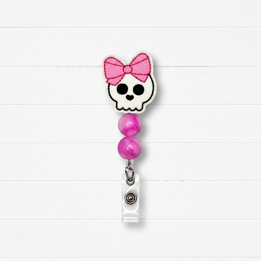 A retractable badge reel with a skull and pink bow design, featuring two pink beads and a silver clip, set against a white wood plank background.