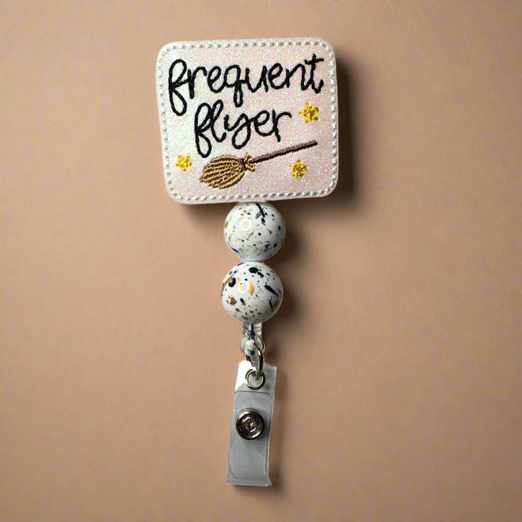 A beaded badge reel with a 'Frequent Flyer' text design, featuring a broom and two black and white beads, attached to a clip.