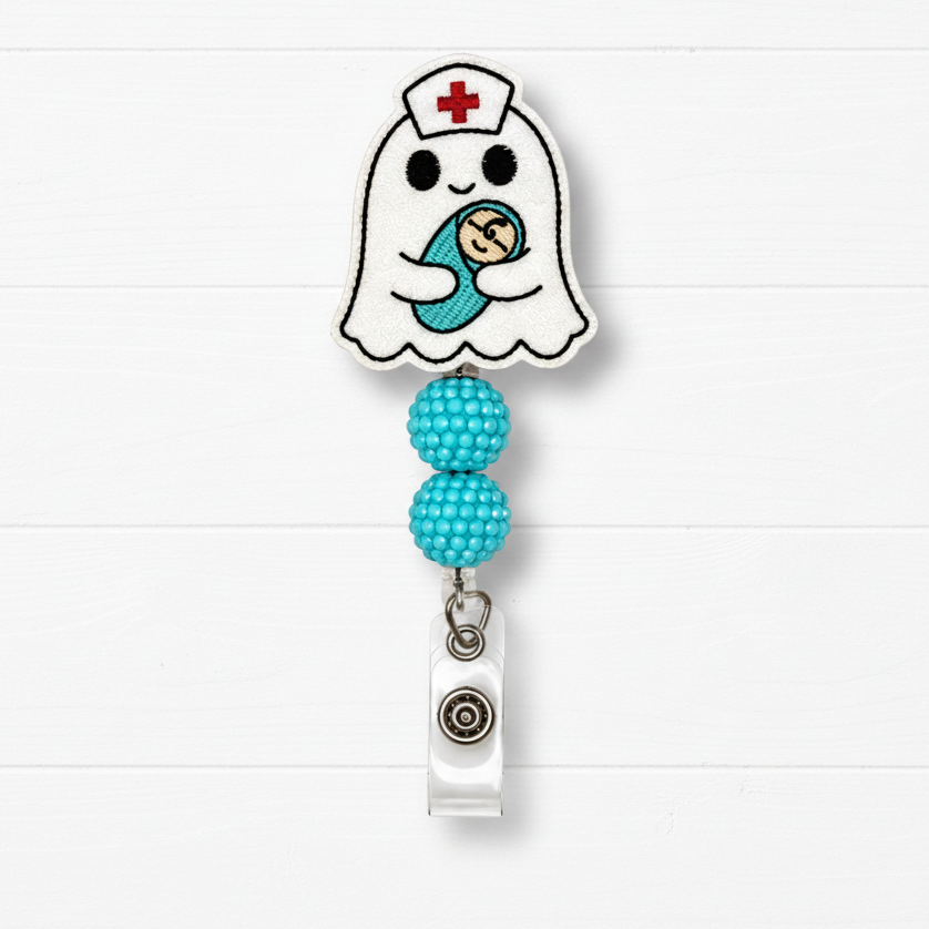 A ghost-shaped nurse badge reel with a cute embroidered baby design, featuring two turquoise beaded loops and a silver retractable clip, set against a white wood plank background.