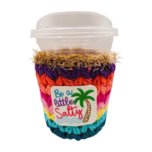 Colorful crocheted coffee cup sleeve with 'Be a little Salty' text and palm tree design on a white background.