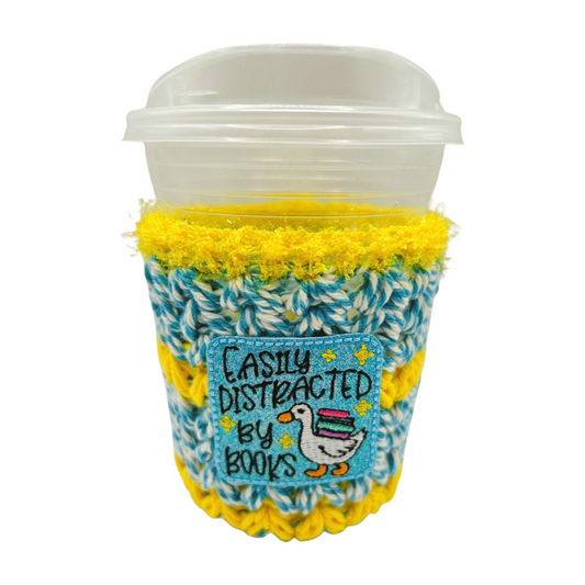Coffee cup with a yellow and blue knit cozy featuring text and a duck illustration.