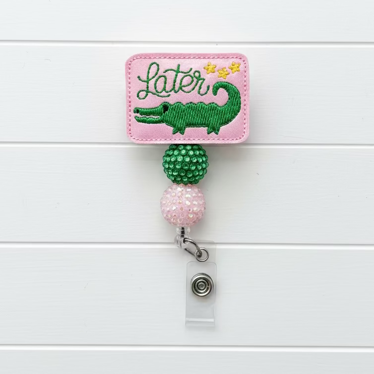 A pink embroidered patch with a green alligator and the word 'Later' in script, attached to a retractable badge reel with green and iridescent beads on a light pink background.