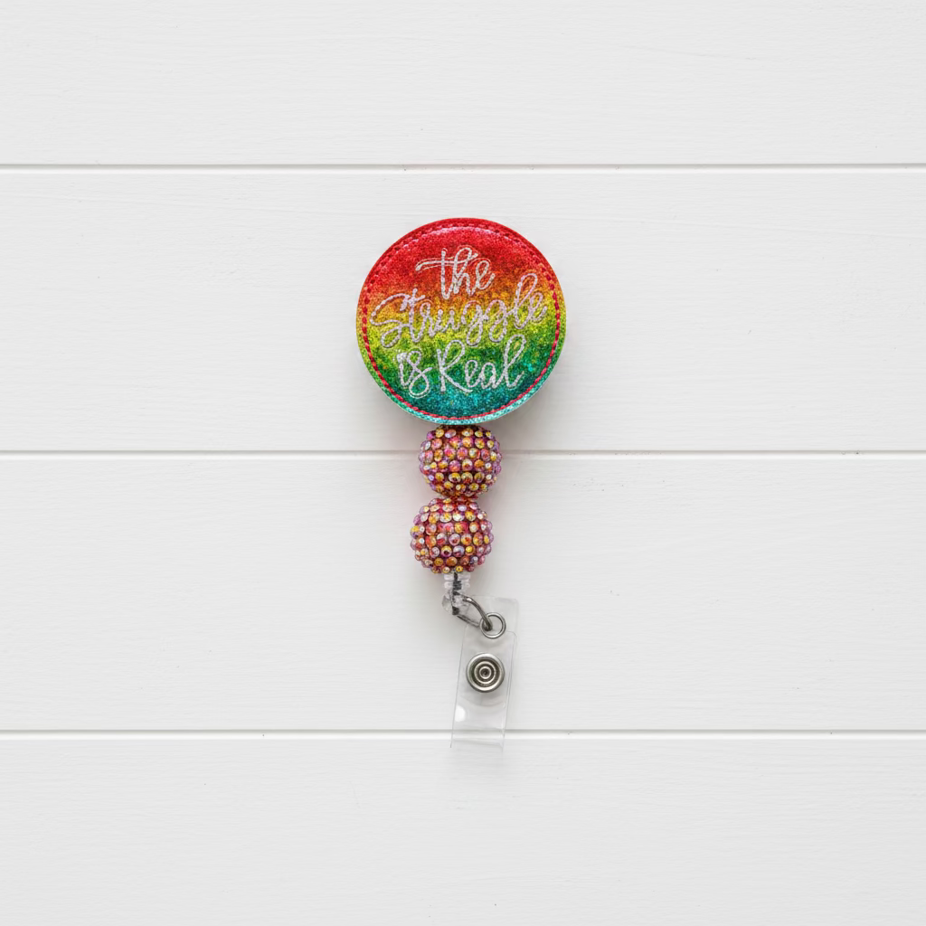 A retractable badge reel with a round top featuring a vibrant rainbow gradient and silver glitter script text, connected by two rhinestone-covered beads to a clear plastic clip with a silver metal snap.