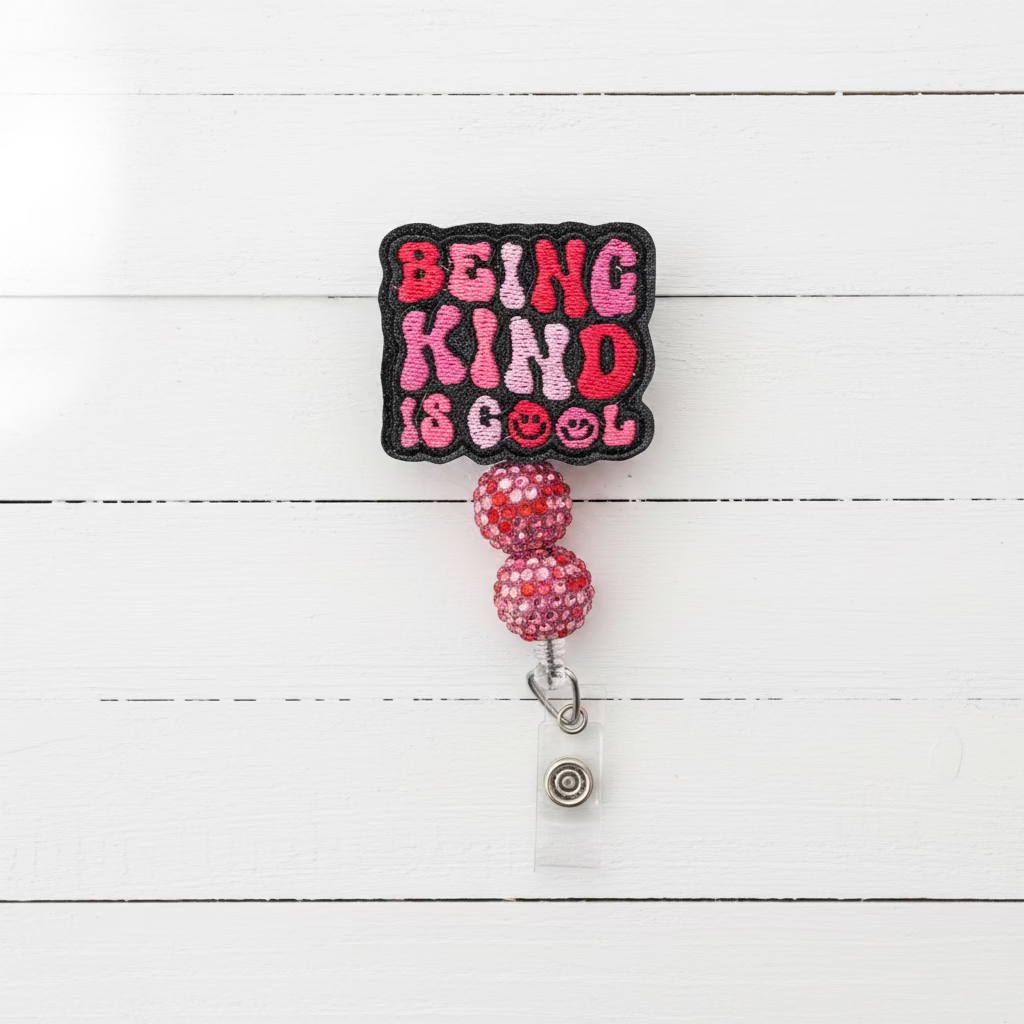 A square embroidered patch with the text 'BEING KIND IS COOL' in pink and red letters on a black background, attached to two pink rhinestone-covered beads and a clear retractable badge reel on a light pink background.