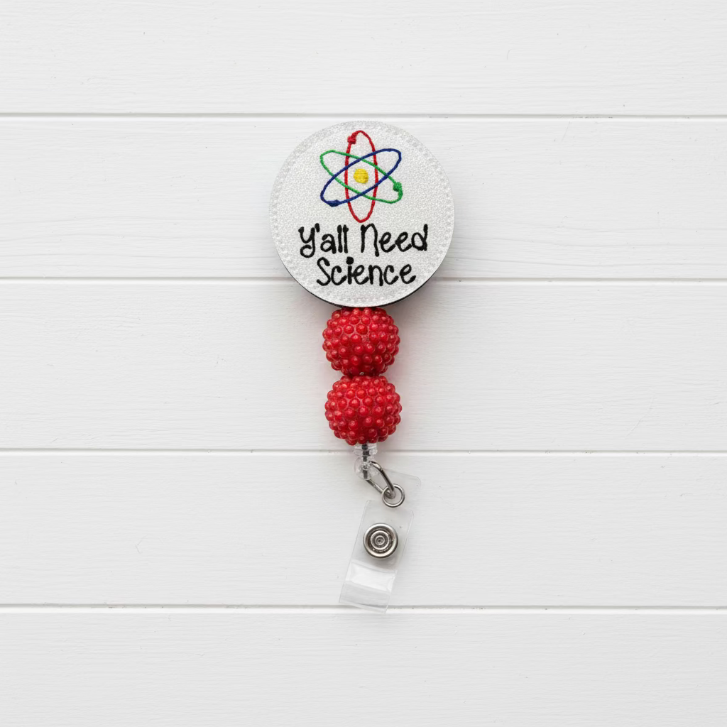 A circular white retractable badge reel featuring the text 'Y'all Need Science' and a colorful atom graphic, with two red beaded loops and a clear plastic clip below, shown on a light pink background.