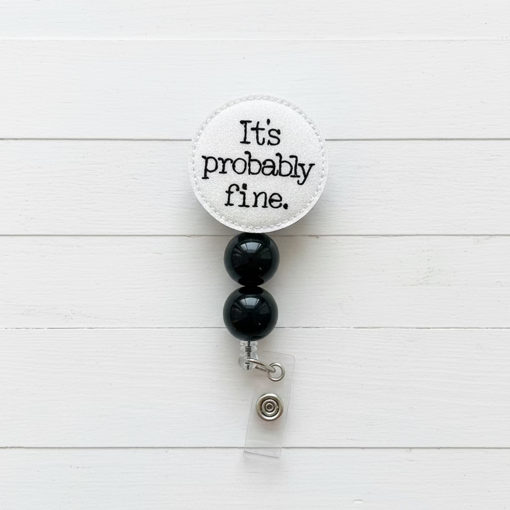 A novelty retractable badge reel with a round off-white fabric top that reads 'It's probably fine.' in black text, attached to two black beads and a silver clasp with a clear plastic backing.