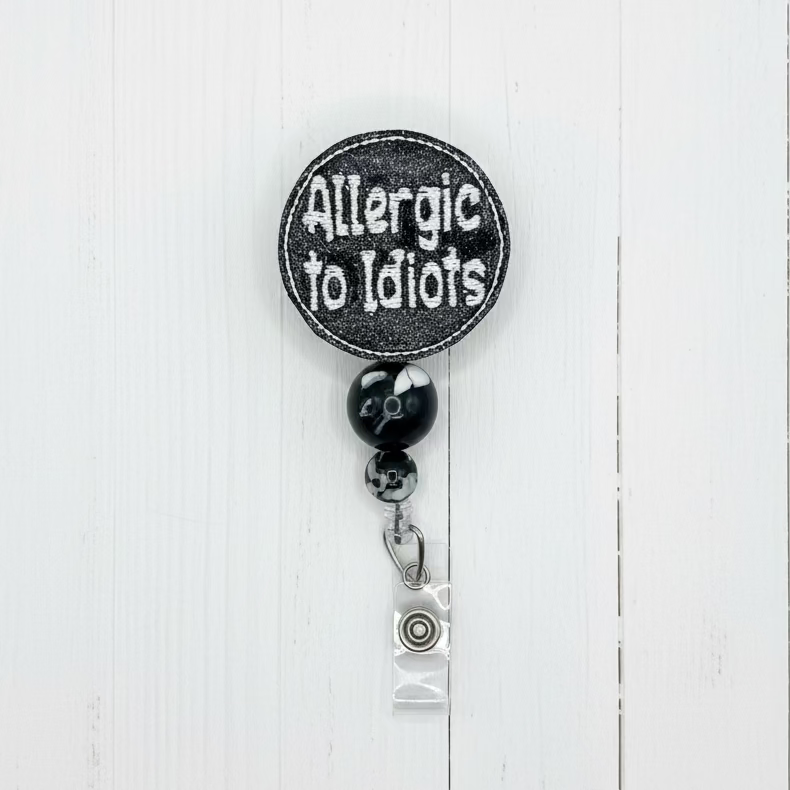 A circular black patch with white embroidered text reading 'Allergic to Idiots' hangs from a black and white marbled bead, a silver lobster clasp, and a clear plastic badge reel on a light pink background.
