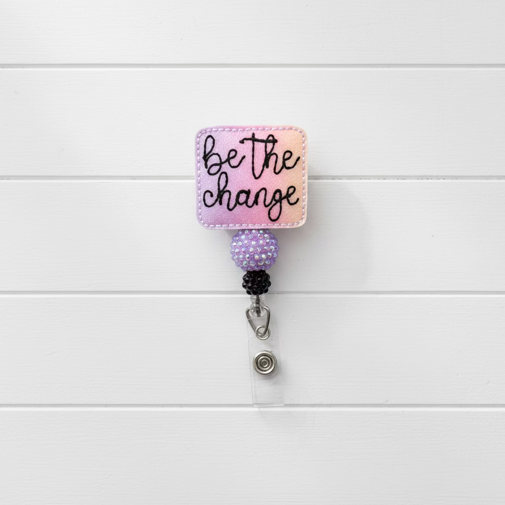 A square embroidered patch with the phrase 'be the change' in black script on a pink and purple gradient background, attached to a retractable badge reel with a clear clip on a light pink background.