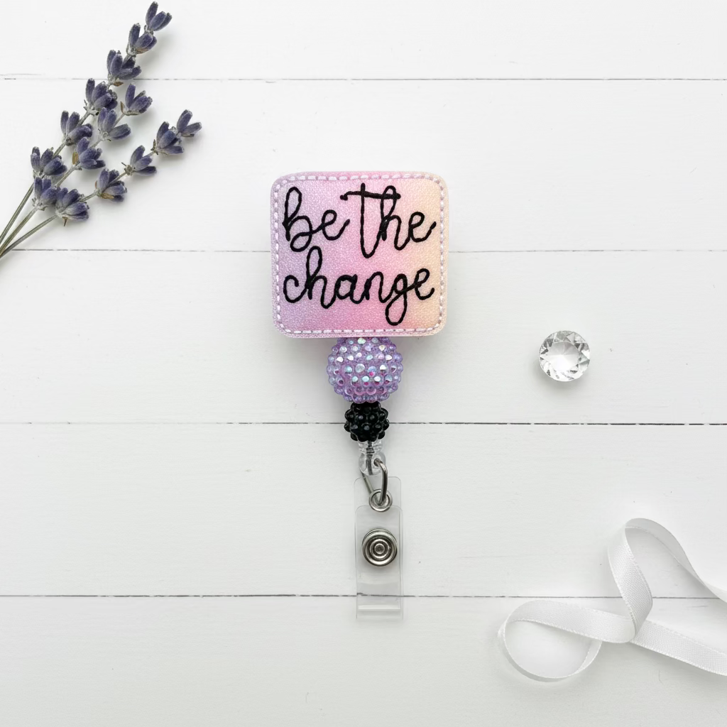 A square embroidered patch with the phrase 'be the change' in black script on a pink and purple gradient background, attached to a retractable badge reel with a clear clip on a light pink background.