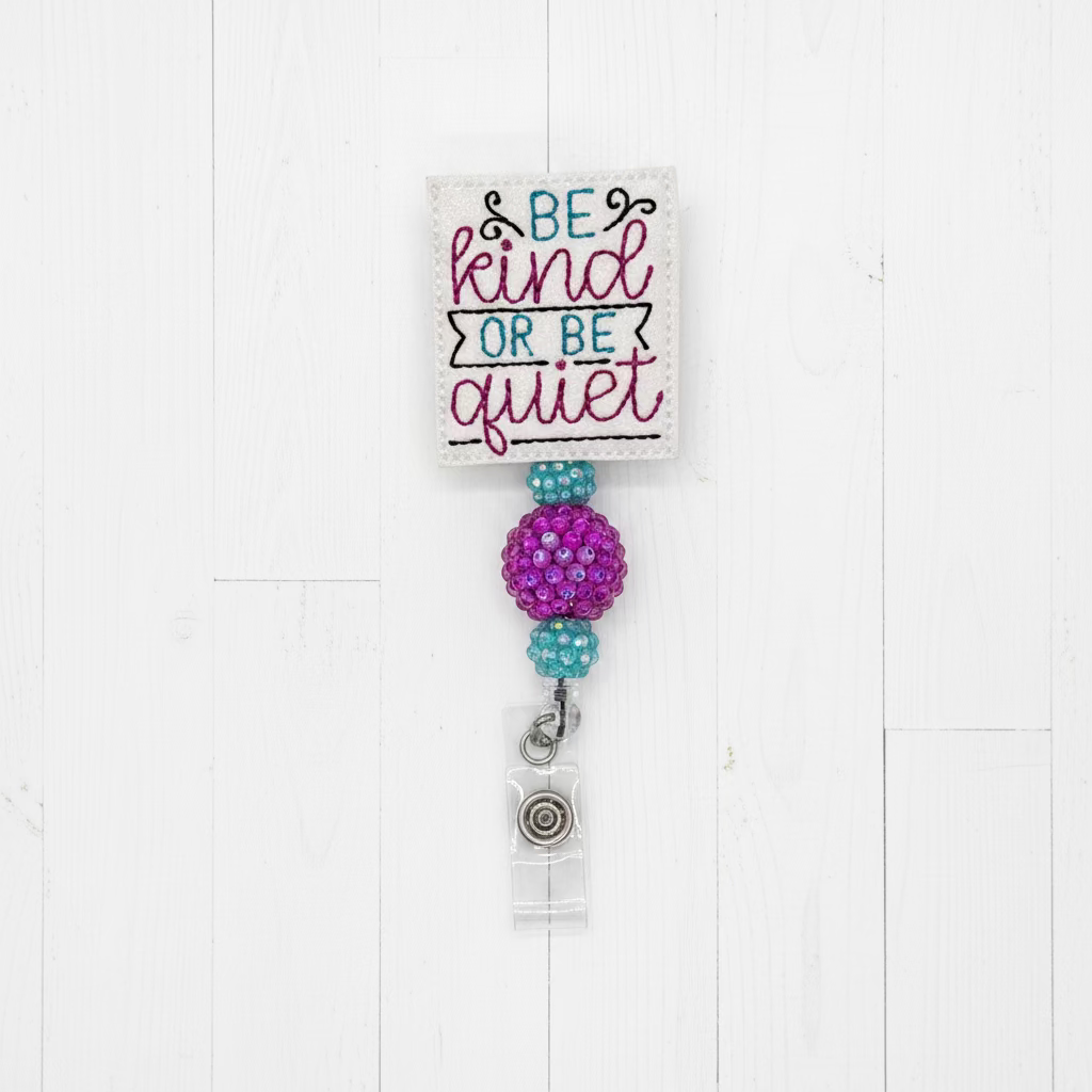 A fabric swatch card with the embroidered text 'BE kind OR BE quiet' in teal and magenta, attached to a purple beaded ball and a silver charm on a light pink background.