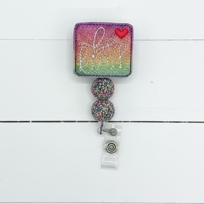 A square glitter badge reel cover with a rainbow gradient background, silver glitter script text, a small red heart, and two dangling glitter-covered beads attached to a clear plastic clip.