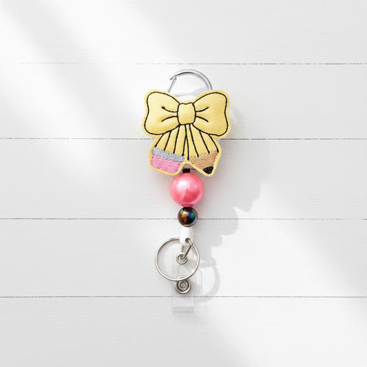 A retractable badge reel keychain featuring a yellow bow design with black outlines, a pink beaded accent, a black bead with a pink logo, and a silver key ring on a light lavender background.