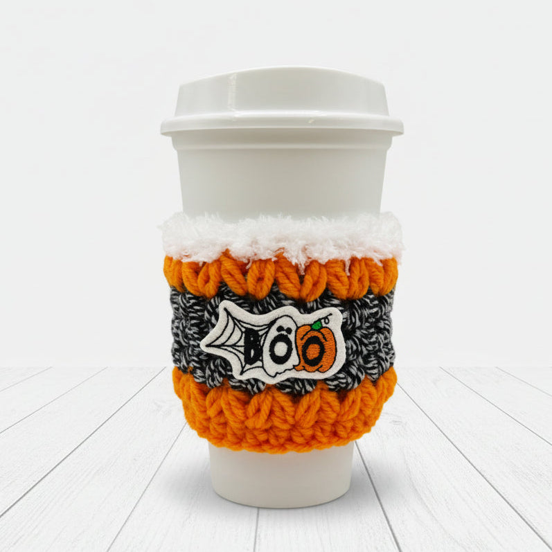A white paper coffee cup with a white lid, wrapped in a knitted Halloween-themed sleeve featuring orange, black, and white colors with a '100' pumpkin design, on a light gray background.