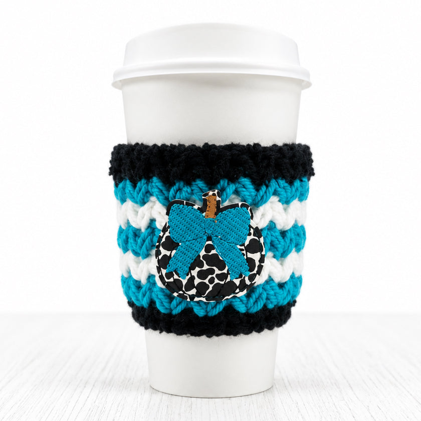 A white paper coffee cup with a white lid, wrapped in a black and white striped knit sleeve featuring a blue bow applique with a leopard print center and a small gold charm.