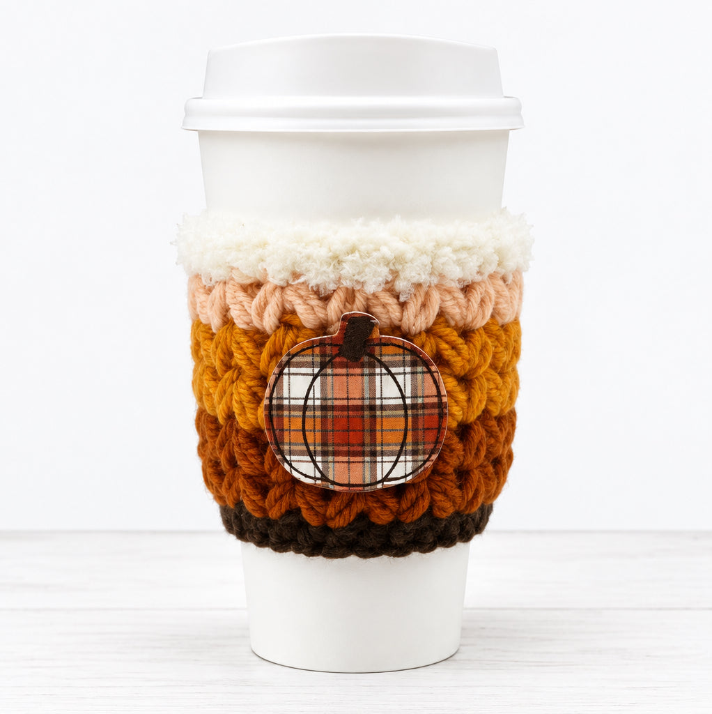 A white paper coffee cup with a white lid, wrapped in a crocheted autumn-themed sleeve featuring stripes of cream, peach, orange, and brown, with a small plaid pumpkin patch detail on the front.