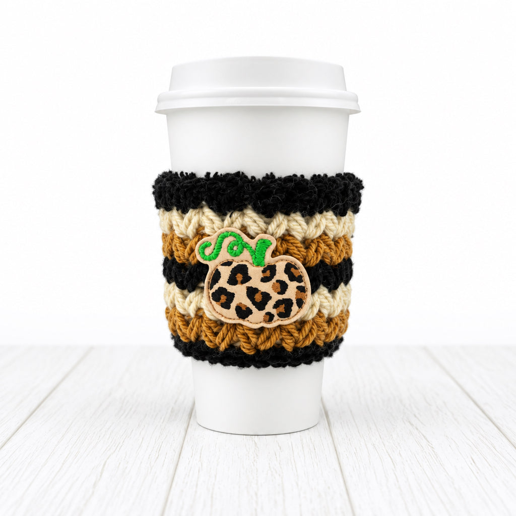 A white paper coffee cup with a white lid, wrapped in a black and tan crochet sleeve featuring a leopard-print pumpkin patch and a green embroidered 'SO' monogram, on a light gray background.