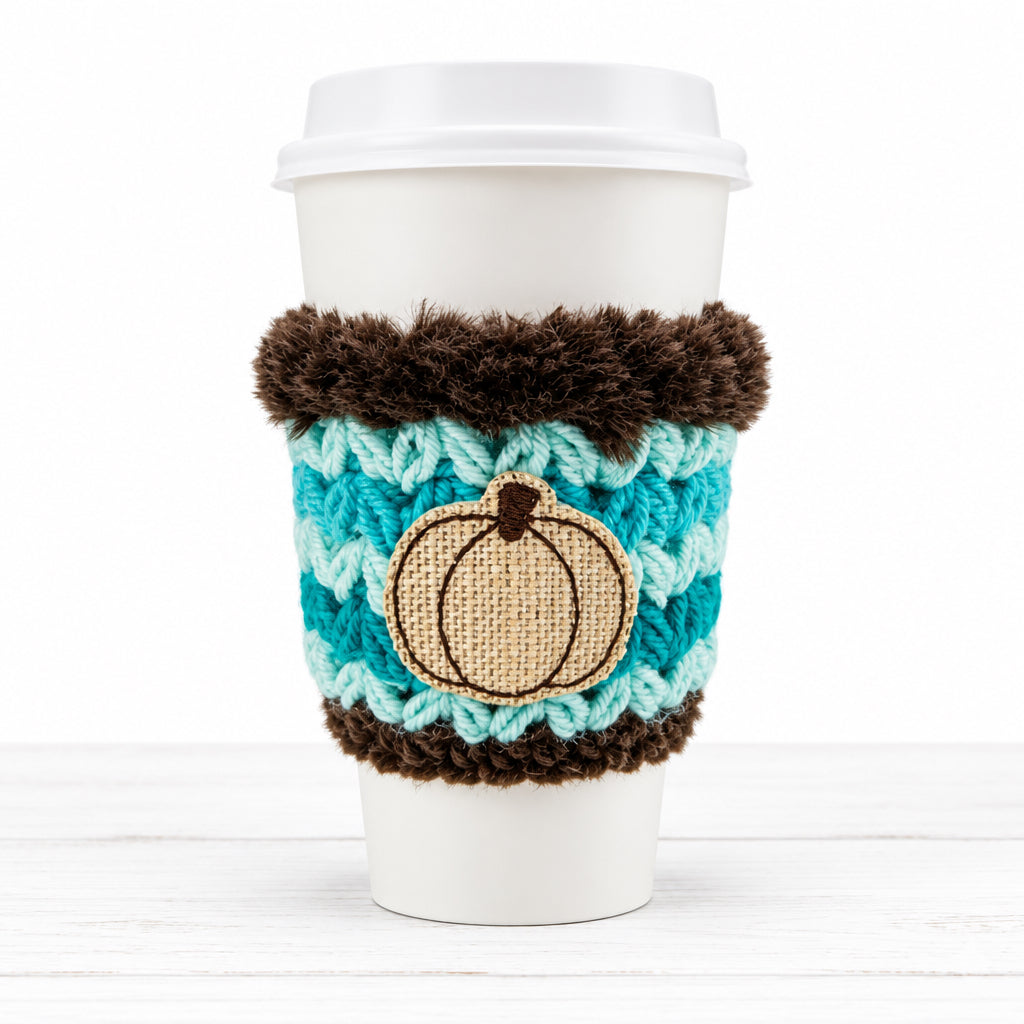 A white disposable coffee cup with a lid, wrapped in a turquoise and brown crocheted sleeve featuring a beige pumpkin applique, on a plain light background.