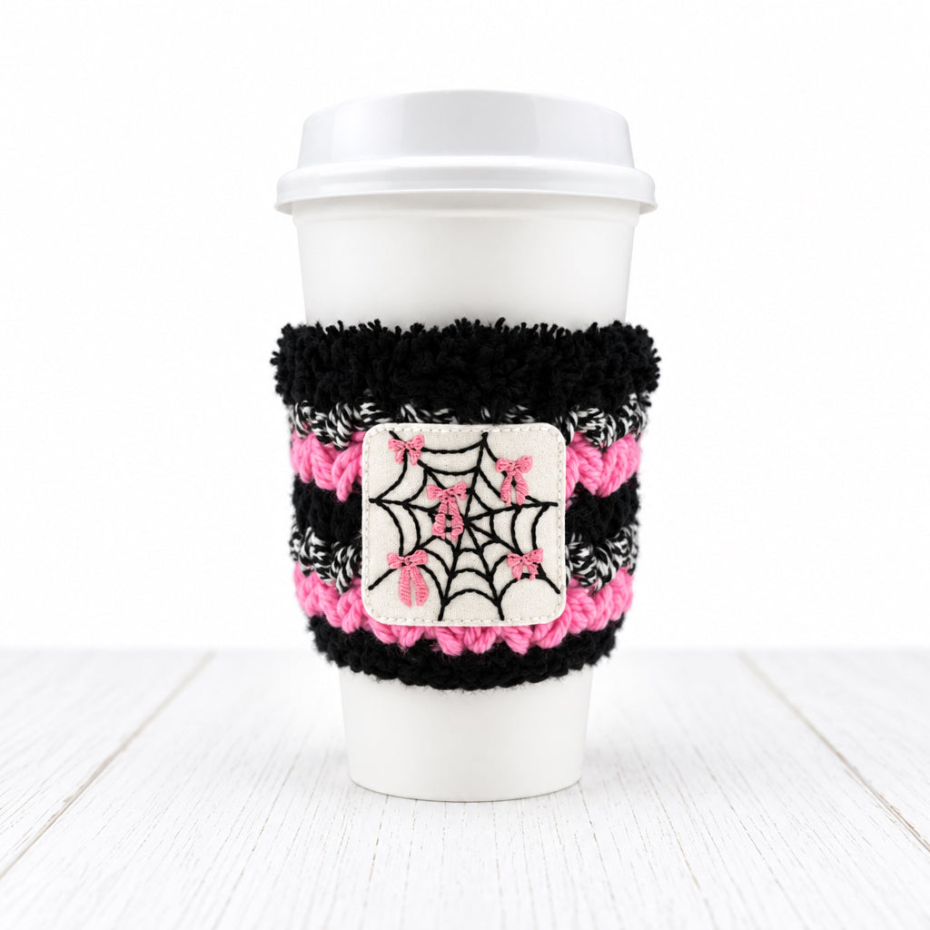 A white disposable coffee cup with a black and pink fuzzy sleeve featuring a white patch with a black spiderweb and pink bows, on a light gray background.