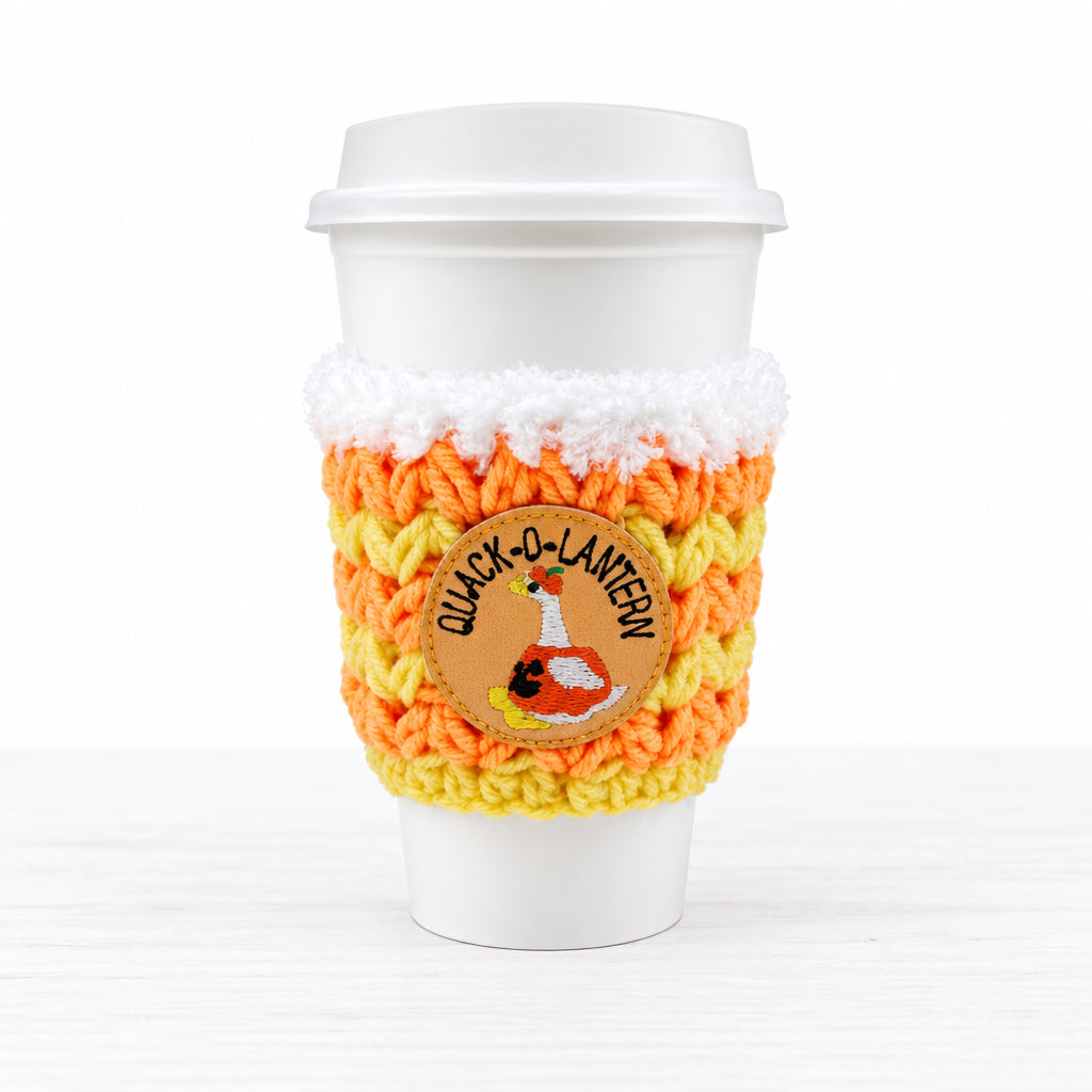 A white disposable coffee cup with a white lid, wrapped in a crocheted sleeve featuring orange, yellow, and white stripes, a faux-fur trim, and a circular 'Quack-O-Lantern' duck patch.