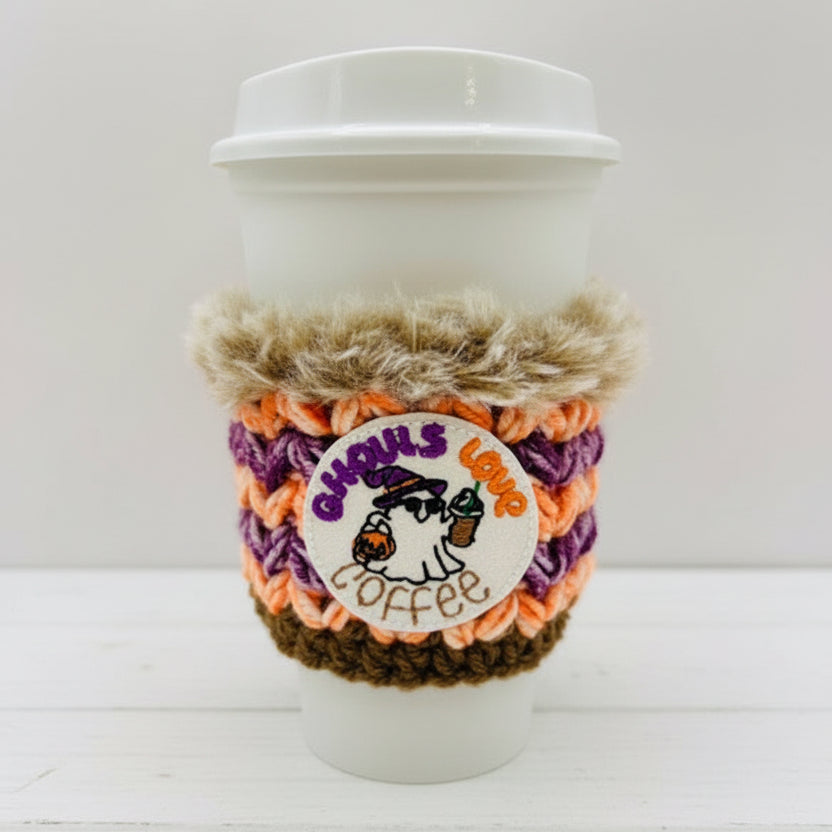 A white paper coffee cup with a lid, wrapped in a handmade crochet Halloween-themed coffee sleeve featuring orange, purple, brown, and beige colors, with a round embroidered 'Ghouls Lamp Coffee' patch.