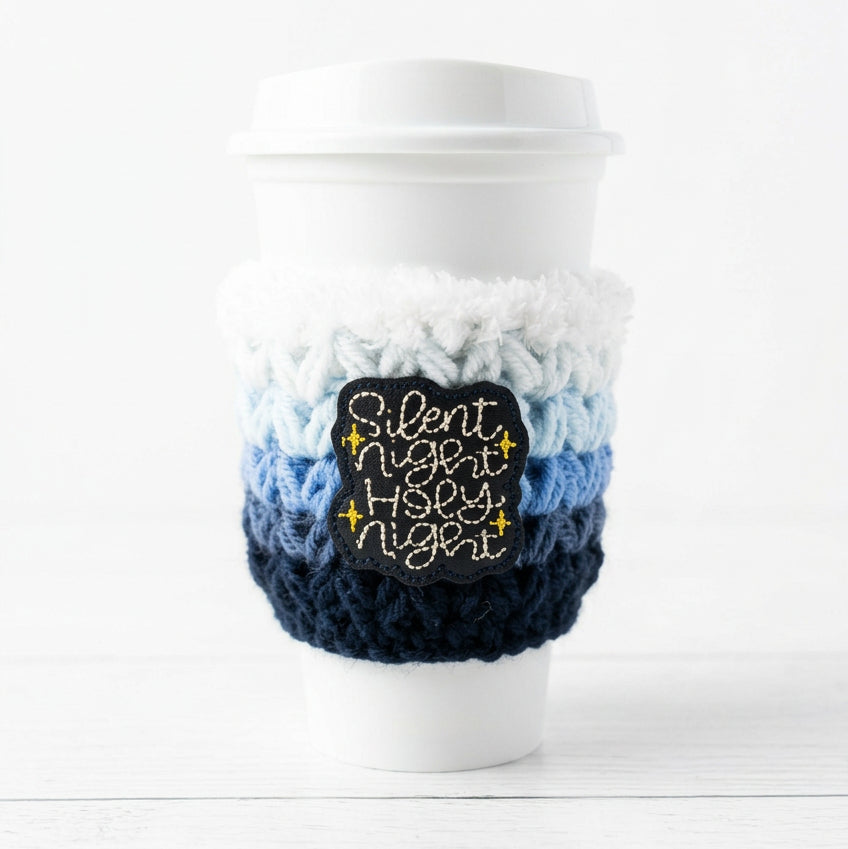 A white disposable coffee cup with a white lid, wrapped in a chunky knit travel mug cozy featuring white, light blue, and navy stripes, with a central embroidered patch reading 'Silent Night Holy Night' in white script with yellow star accents.