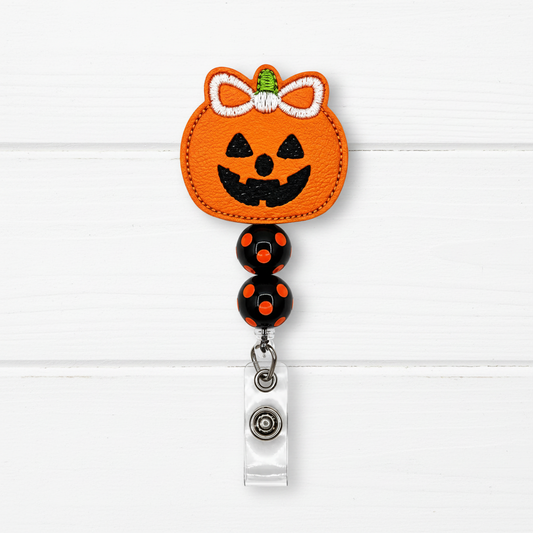 A Halloween-themed retractable badge reel featuring an orange pumpkin patch with a black jack-o'-lantern face, white bow, and green stem, attached to black beads with orange dots and a silver clip on a white wood plank background.
