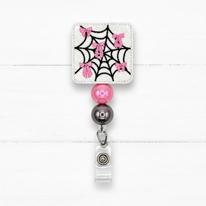 A square embroidered patch with a black spiderweb and pink glitter bows on a white fabric background, attached to a retractable badge reel with pink and dark gray beads.