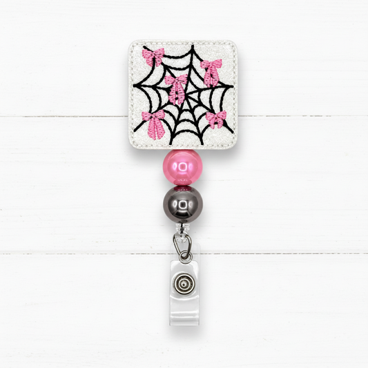 A square embroidered patch with a black spiderweb and pink glitter bows on a white fabric background, attached to a retractable badge reel with pink and dark gray beads.