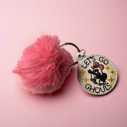 A pink pom pom keychain with a circular keyring and a charm featuring the text 'LETS GO GHOULS' on a pink background.